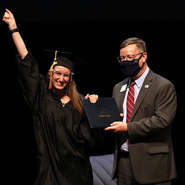 A GED graduate wearing a cap, gown and face shield receives her diploma while smiling at the camera and raising a fist in the air